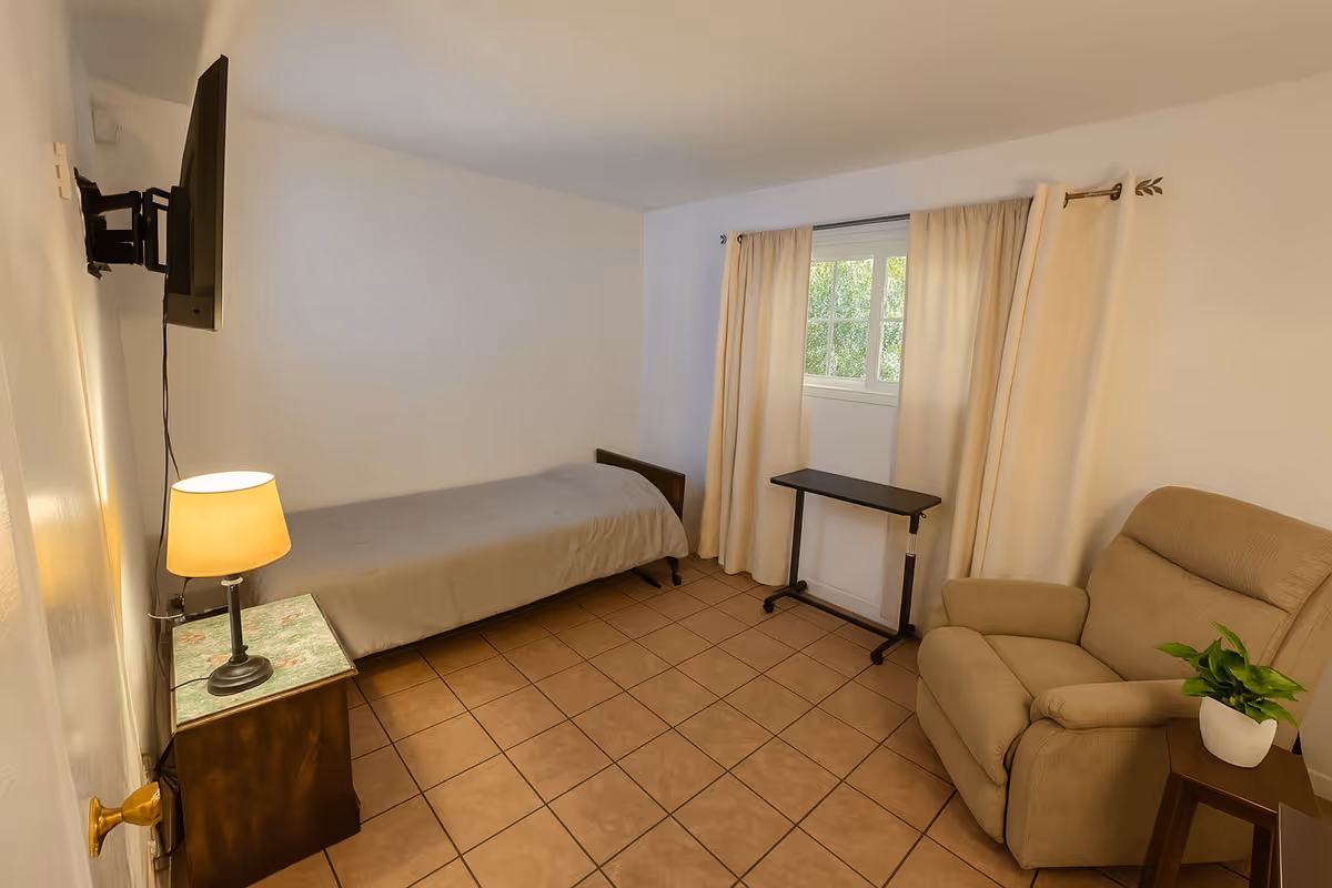 Small furnished bedroom with a single bed, recliner, wall-mounted TV, side table with a lamp, and a window with curtains.