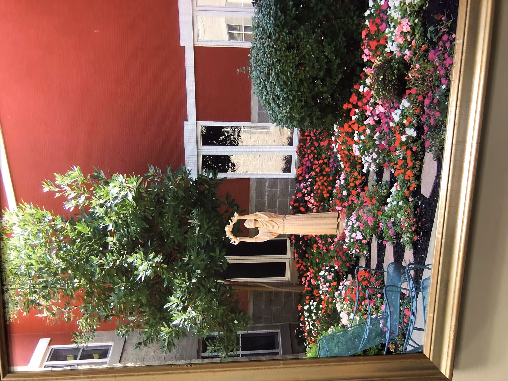 A vibrant garden area with a variety of colorful flowers and a green leafy tree. There is a statue of a person holding a wreath in the center of the flower bed. Behind the garden is a red building with white window frames. In the foreground, there are green metal chairs and a table, suggesting a seating area within the garden.