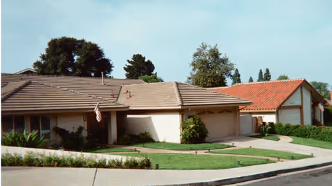 Single-story residential buildings with beige walls and brown and red tiled roofs, surrounded by green lawns and trees under a clear sky.