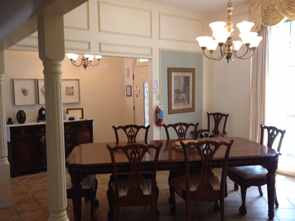A dining room with a large wooden dining table surrounded by six matching wooden chairs. The room features a chandelier with multiple lights hanging above the table, a framed picture on the wall, and a window with curtains allowing natural light to enter. In the background, there is a sideboard with decorative items and framed pictures on the wall.