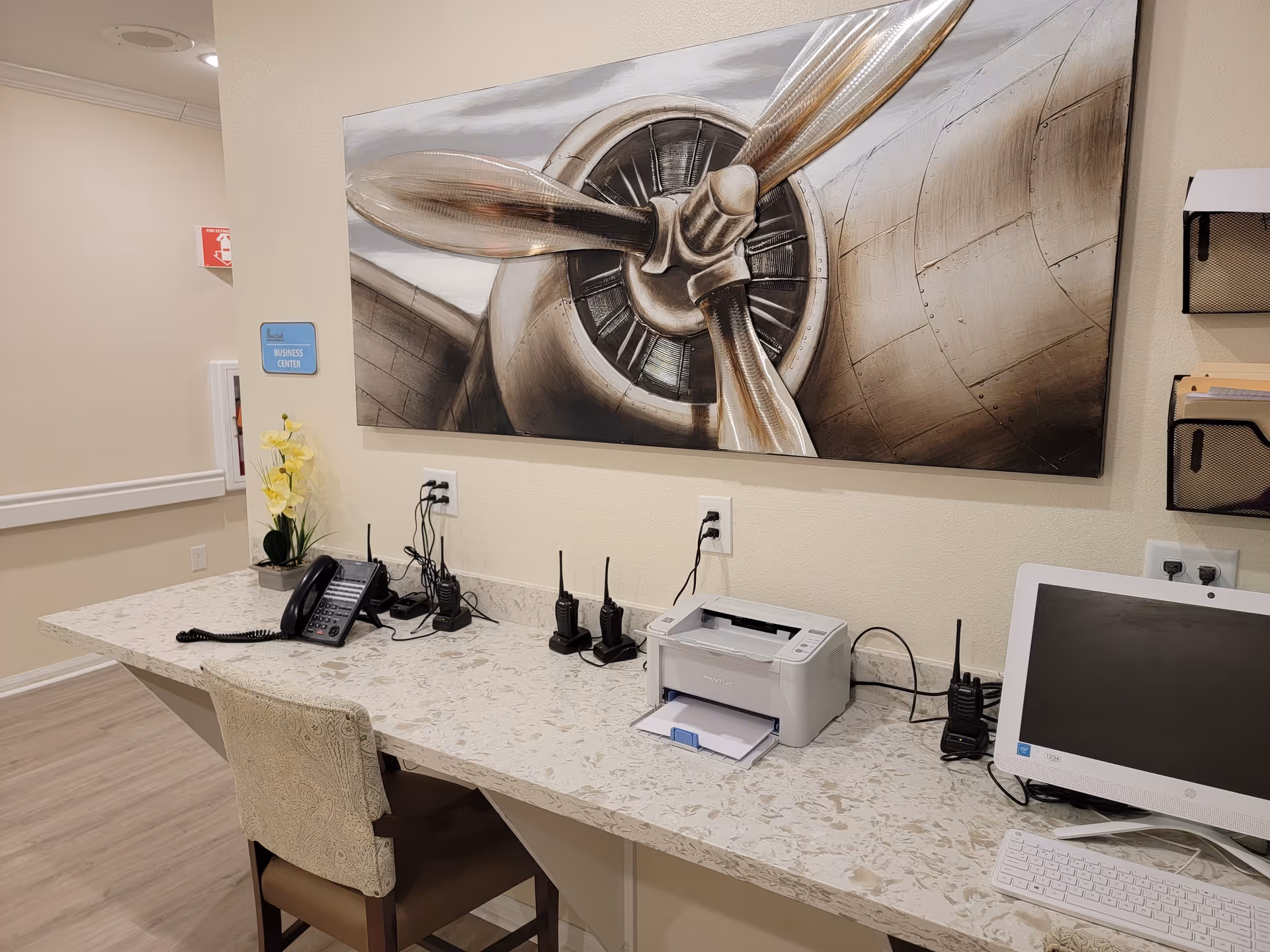 A business center area with a long marble countertop holding a telephone, several walkie-talkies, a printer, and a computer monitor with keyboard. A chair is placed in front of the counter. On the wall above the counter is a large painting of an airplane propeller. A small sign on the wall reads 'Business Center'.