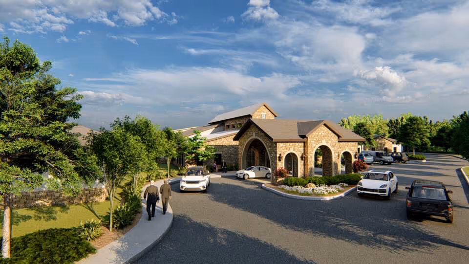 Exterior view of a senior living facility with a stone building featuring arched entrances, surrounded by trees and landscaping. Several cars are parked in the driveway and parking area, and two people are walking on the sidewalk towards the building under a partly cloudy sky.