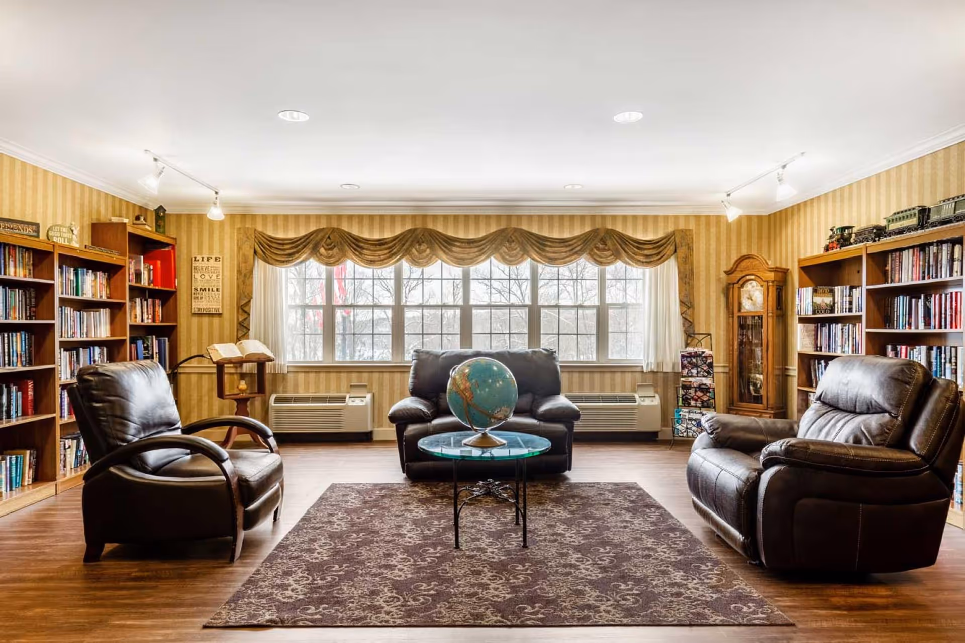 A cozy senior living common area with three dark leather armchairs arranged around a glass coffee table holding a globe. The room features large windows with decorative valances, wooden bookshelves filled with books on either side, a grandfather clock, and a patterned area rug on wooden flooring.