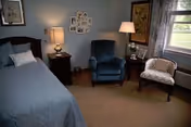 A cozy bedroom with a single bed covered in a blue bedspread, a dark wooden nightstand with a lamp and decorative items, a blue upholstered armchair, a white patterned chair, a window with curtains, and framed artwork on the walls.