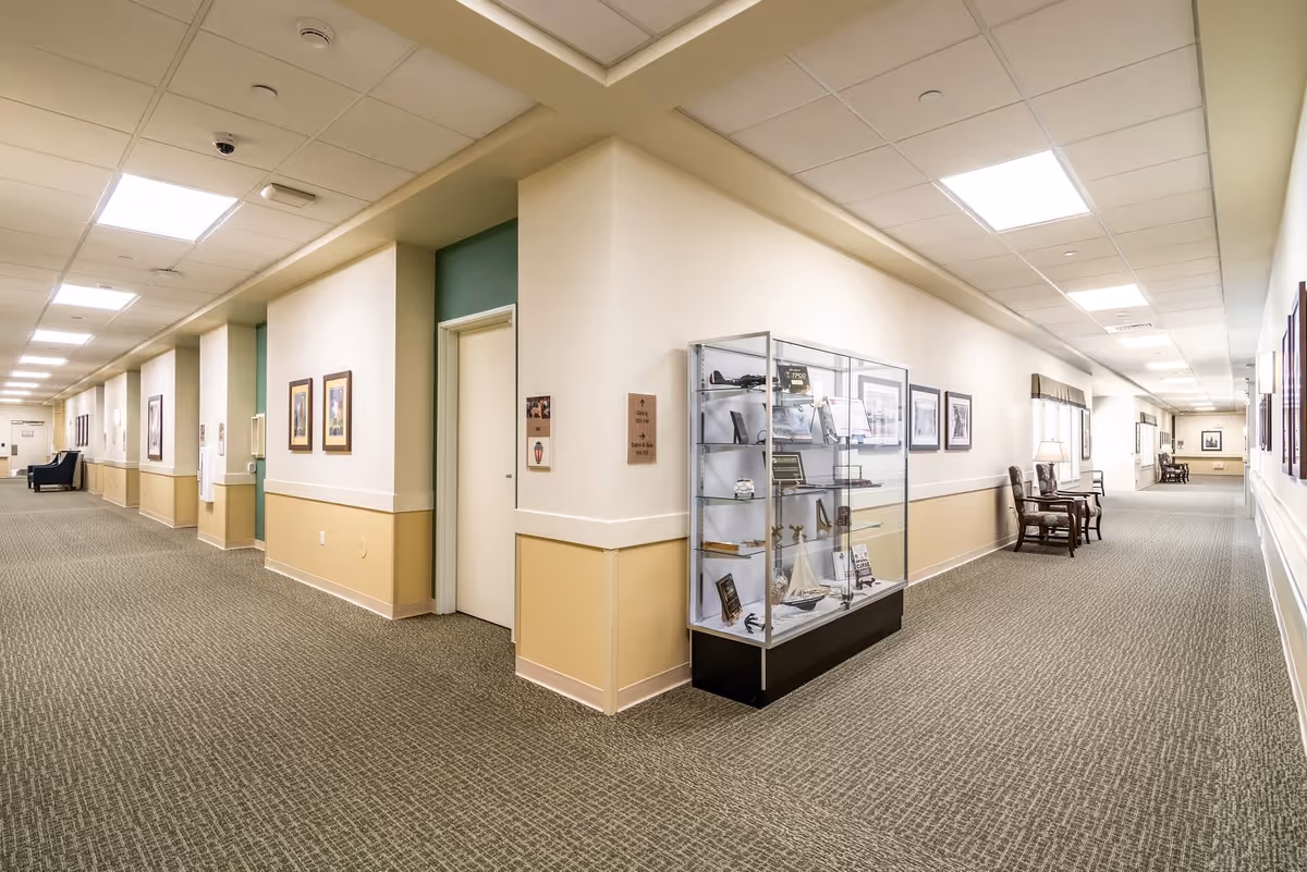A wide, well-lit hallway in a senior living facility with beige and light green walls, carpeted floor, and ceiling lights. The hallway features framed pictures on the walls, a glass display case with various items, and several chairs along the walls for seating.