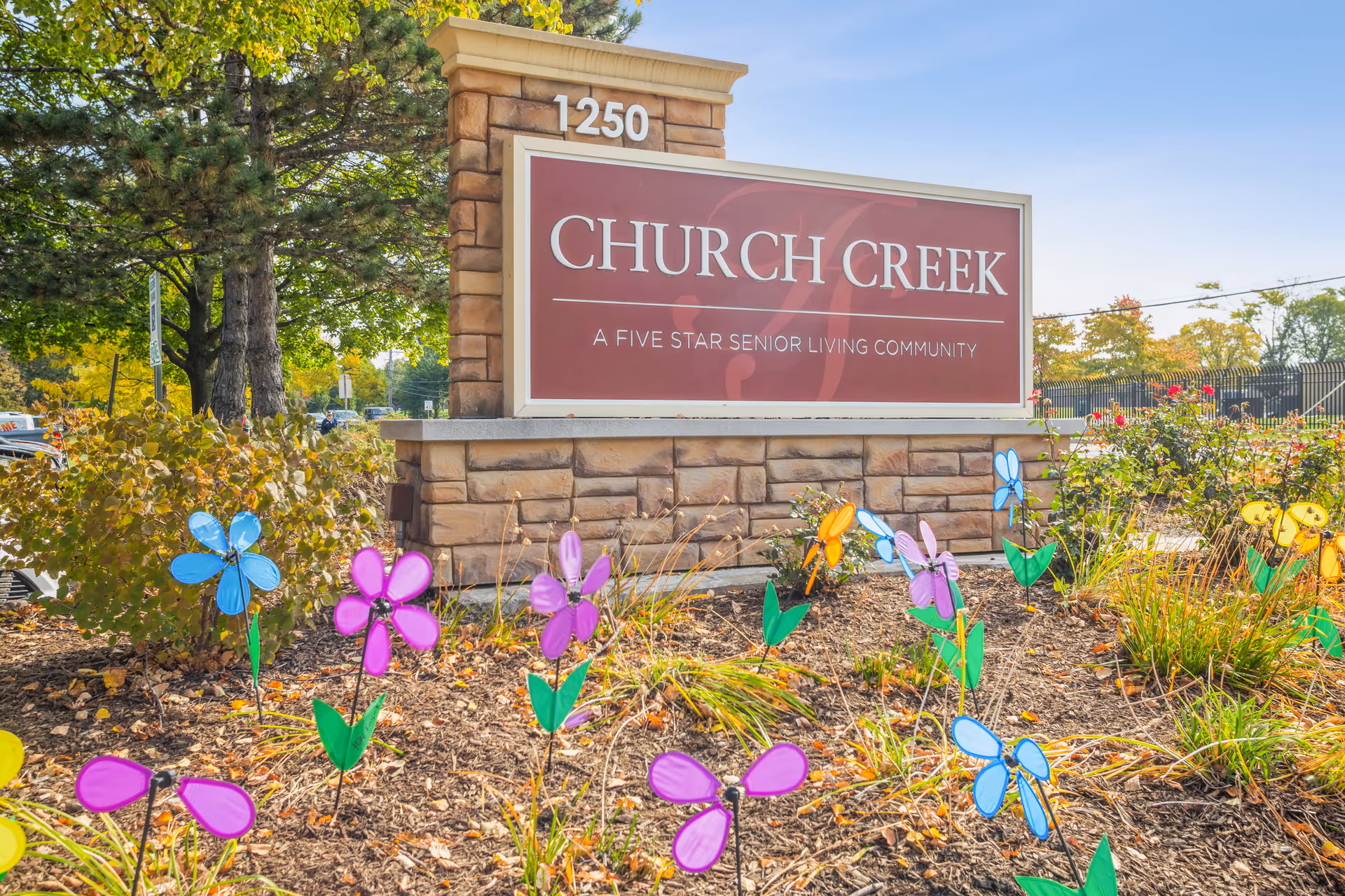 Outdoor view of a stone sign for Church Creek, a five star senior living community, with colorful decorative pinwheel flowers planted in the garden bed in front of the sign and trees in the background.