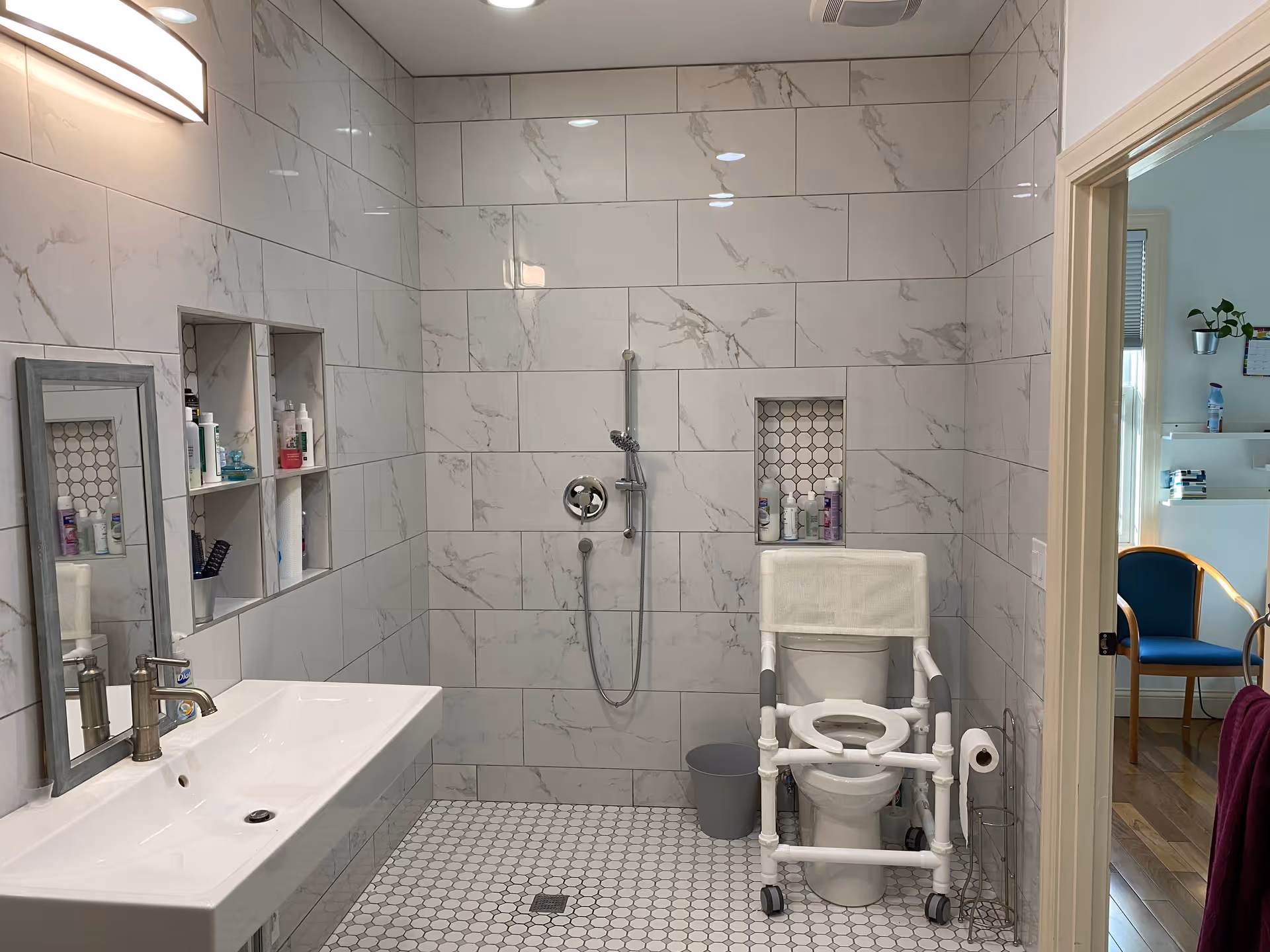 A clean, accessible bathroom with white marble-patterned tiles on the walls and small hexagonal tiles on the floor. The bathroom features a wall-mounted sink with a mirror above it, built-in shelves holding toiletries, a walk-in shower with a handheld showerhead, and a toilet equipped with a white support frame for assistance. A doorway leads to an adjacent room with a blue chair and wooden floor.