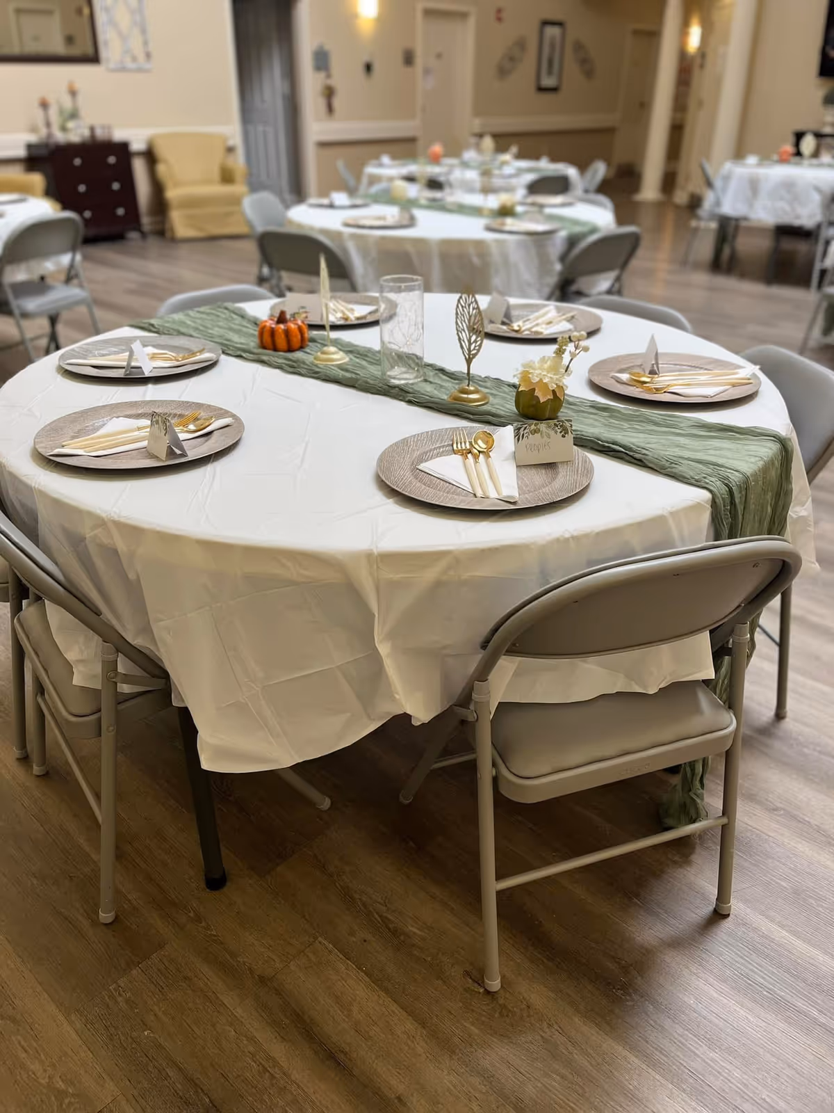 A round dining table set with a white tablecloth and a green table runner, decorated with small pumpkins and gold leaf ornaments. Each place setting has a woven placemat, white napkin, gold utensils, and a name card. The room has wooden flooring and other similarly set tables in the background.