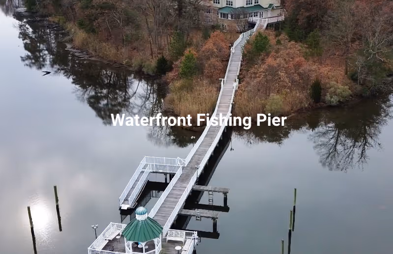 A long wooden fishing pier extending over calm water with a small gazebo at the end, surrounded by trees with autumn foliage and a building in the background.