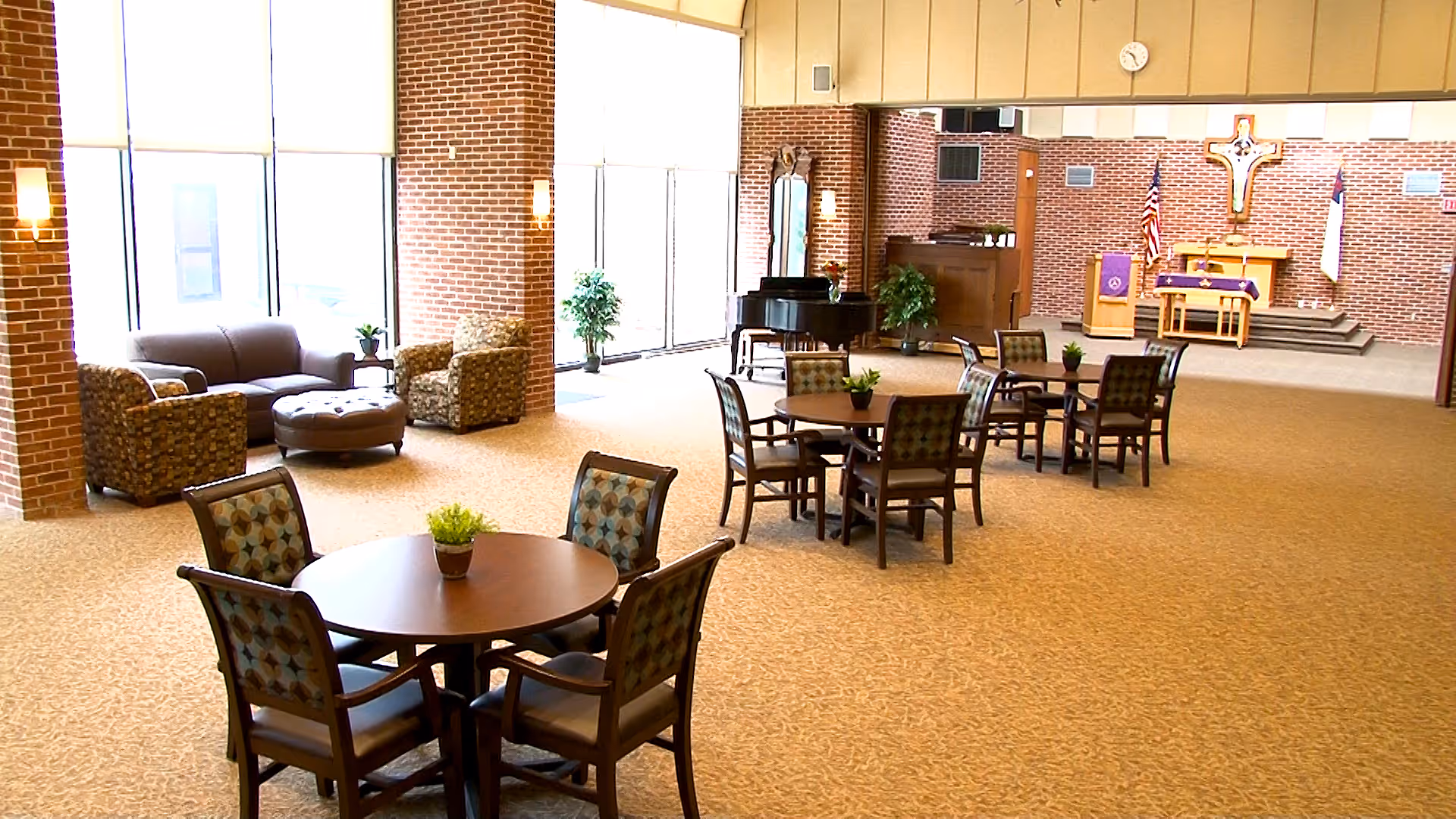 A spacious senior living common area with multiple round tables and chairs arranged for socializing or activities. There is a seating area with a sofa, two armchairs, and an ottoman near large windows letting in natural light. In the background, there is a piano and a small stage with religious symbols including a cross and flags.