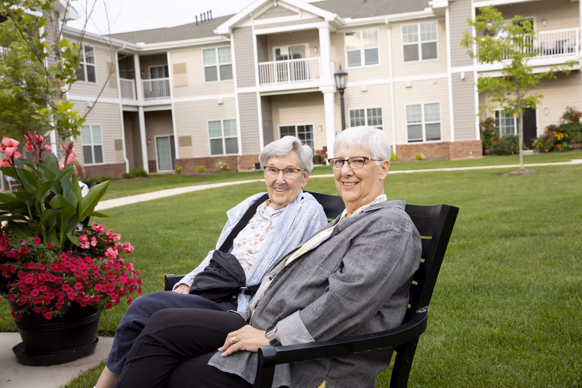 Two elderly women sitting on a black bench outside on a grassy lawn in front of a beige multi-story residential building with balconies and windows. There is a large flower pot with vibrant pink and red flowers to the left of the bench.