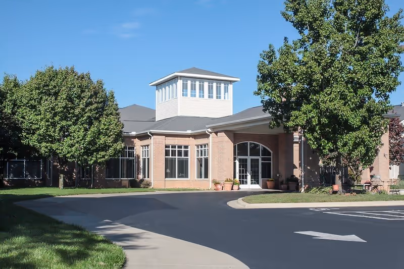 Exterior view of a single-story brick building with large windows and a central entrance under a covered porch. The building has a distinctive tower-like structure with multiple windows on top. There are trees and potted plants around the entrance, and a paved driveway with a sidewalk leading to the door under a clear blue sky.