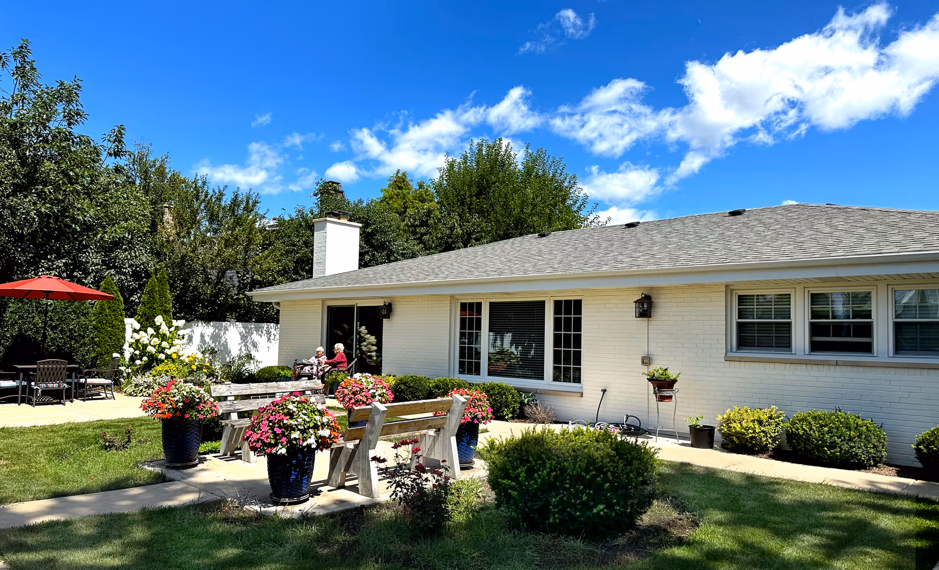 Outdoor garden area of a senior living facility with benches, flower pots, and a patio umbrella. Two elderly people are sitting and conversing near the building under a bright blue sky with scattered clouds.