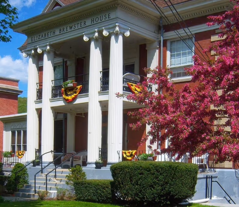 Front exterior view of Elizabeth Brewster House, a red brick building with white columns and a balcony decorated with yellow and red bunting. There are green bushes and a tree with pink blossoms in front of the building under a blue sky.