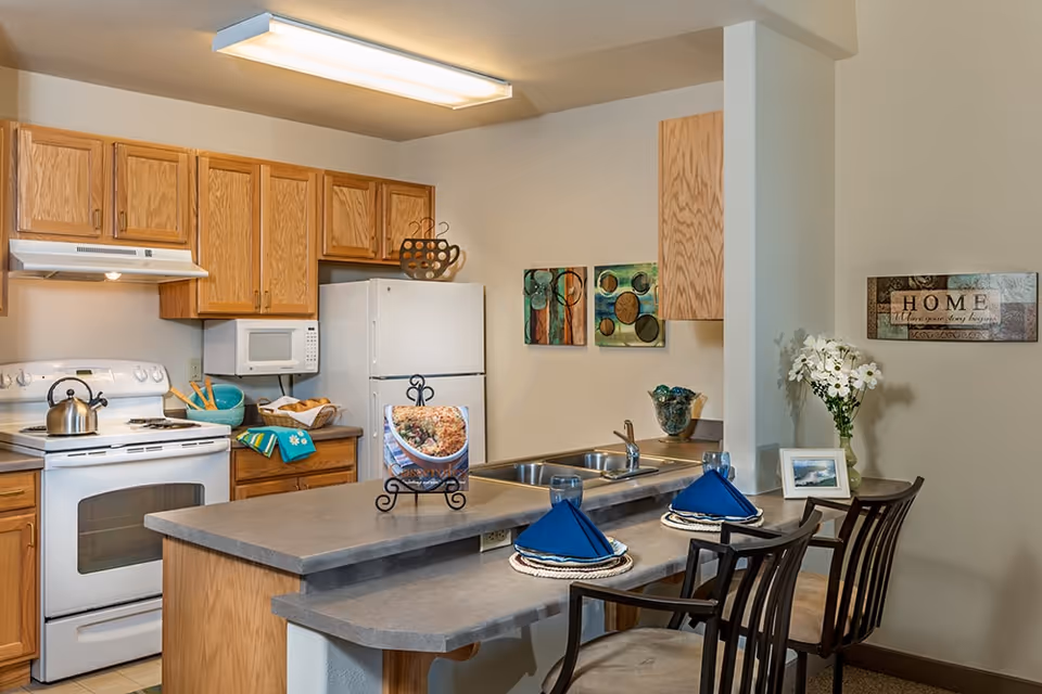 A cozy kitchen area with wooden cabinets, a white stove with a kettle, a microwave, and a refrigerator. There is a countertop with two place settings featuring blue napkins and glasses, two chairs, and decorative items including wall art, a vase with flowers, and a framed photo.