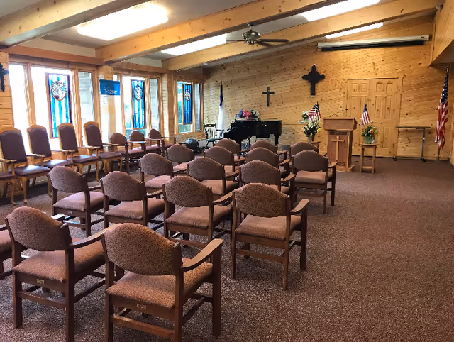 Interior view of a chapel or meeting room with rows of brown cushioned chairs arranged facing a wooden podium and a black grand piano. The room has wooden walls, stained glass windows on the left, a ceiling fan, and flags including an American flag and a Christian flag.