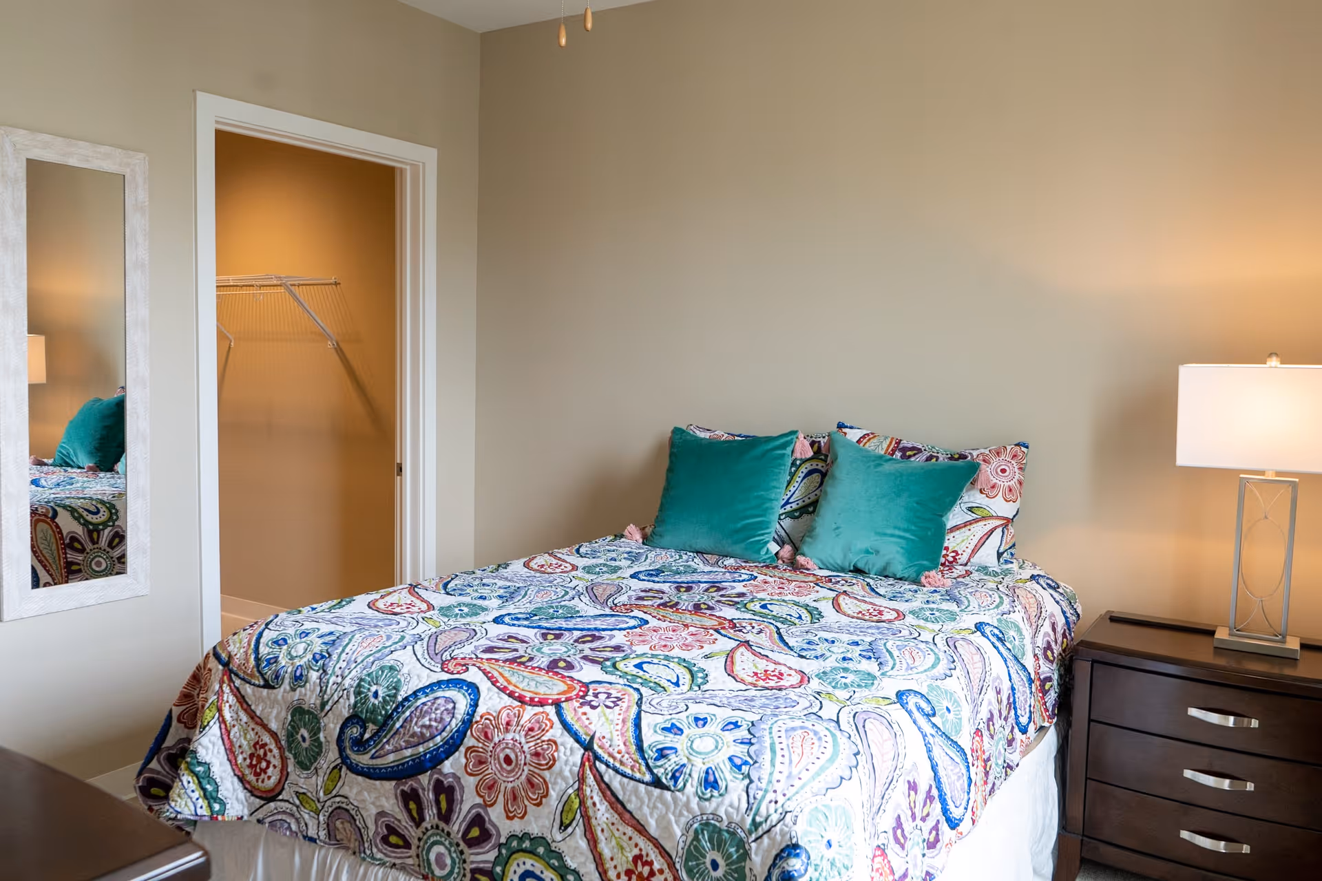 A bedroom with a bed covered in a colorful paisley-patterned bedspread and two teal pillows. There is a dark wooden nightstand with a lamp on the right side of the bed. A mirror is mounted on the wall to the left, and an open door reveals a walk-in closet with wire shelving.