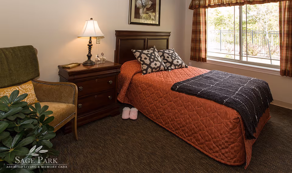 A cozy bedroom with a single bed covered in a rust-colored quilt and two patterned pillows. A dark wooden nightstand with a lamp and decorative items sits next to the bed. A comfortable armchair with a green throw and a yellow cushion is positioned near a leafy green plant. A window with plaid curtains lets in natural light, and a framed picture hangs on the wall above the bed. Pink slippers are placed on the floor beside the bed.