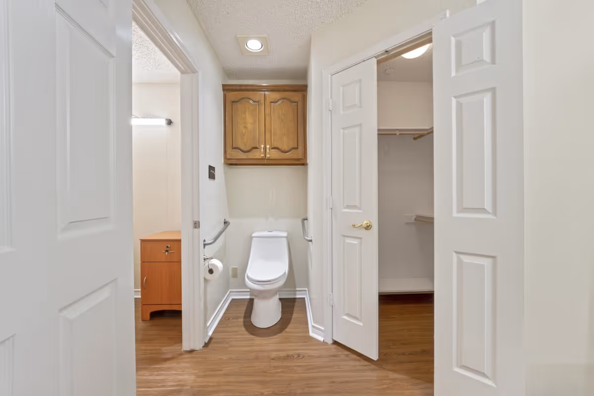 A small bathroom area with a white toilet centered against the wall, wooden cabinet mounted above it, and grab bars on both sides. To the left, there is a doorway leading to a sink with a wooden vanity and a mirror with a light above it. To the right, there is an open door revealing a walk-in closet with wooden flooring and shelving.