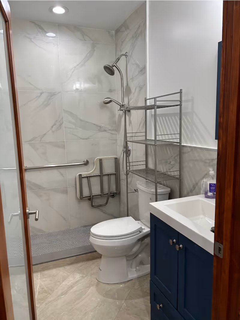 A modern bathroom with a walk-in shower featuring a handheld and overhead showerhead, a foldable shower seat, and a grab bar. There is a white toilet next to a metal shelving unit, and a blue vanity with a white sink and a soap dispenser on top. The walls are tiled with large marble-patterned tiles and the floor has beige tiles.