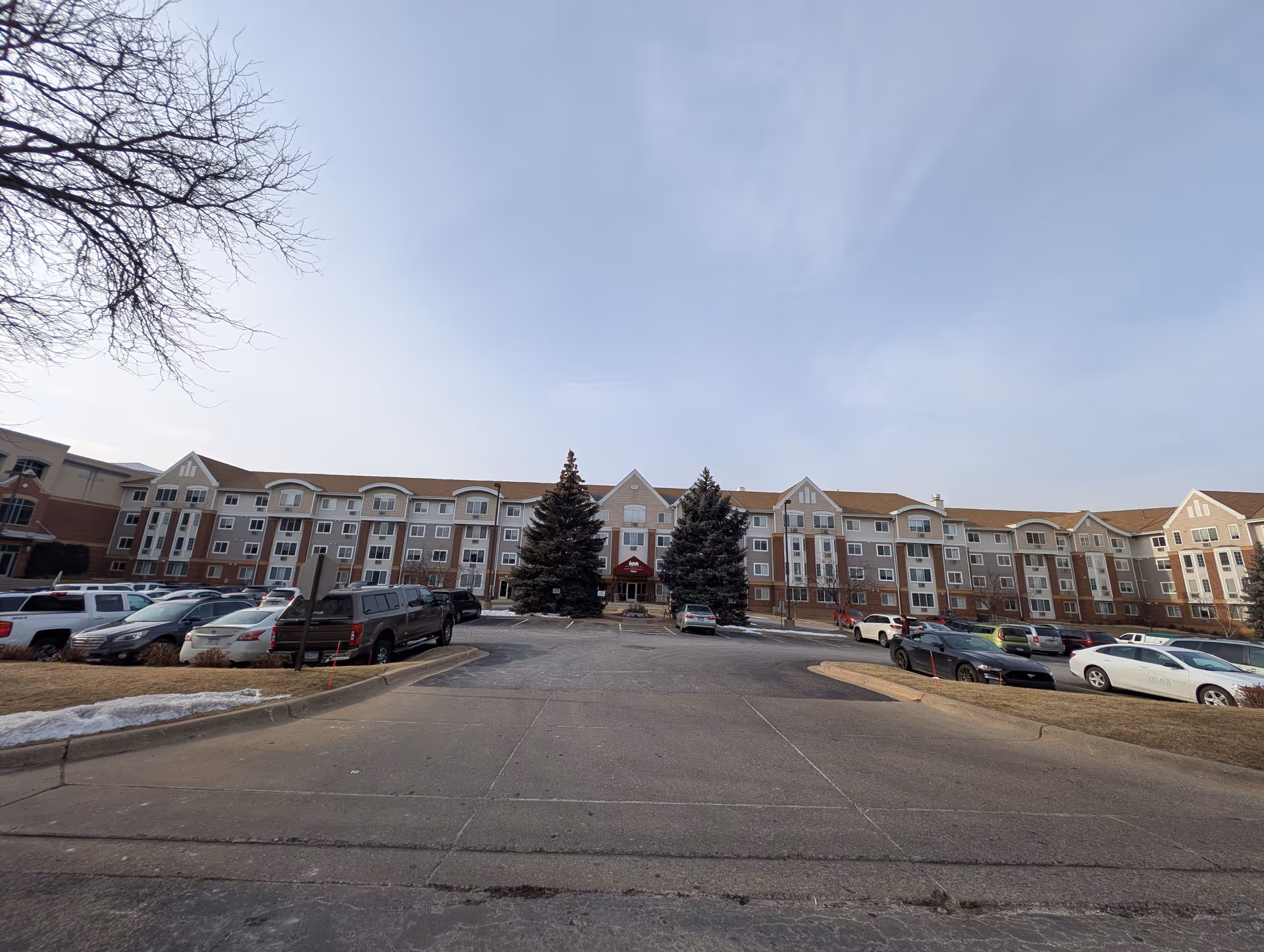 Wide exterior view of a multi-story senior living facility named Mainstreet Village with a large parking lot in front, several parked cars, and two large evergreen trees flanking the main entrance under a clear sky.