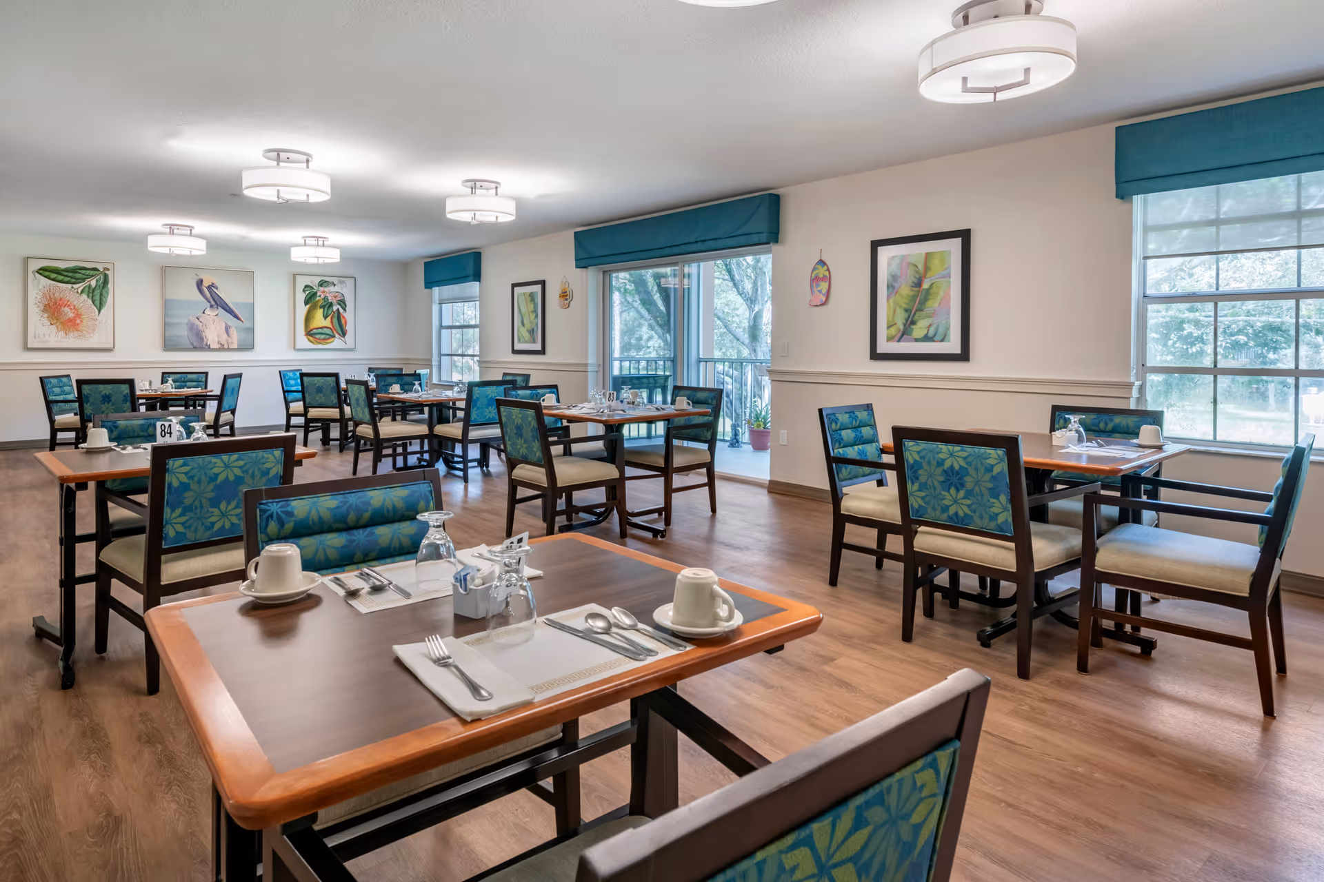 A bright and clean dining room with several wooden tables and chairs featuring blue and green patterned upholstery. Each table is set with white cups, glasses, utensils, and napkins. The room has large windows with blue valances, allowing natural light to fill the space. The walls are decorated with framed botanical and bird artwork. The floor is wood, and modern ceiling lights illuminate the room.