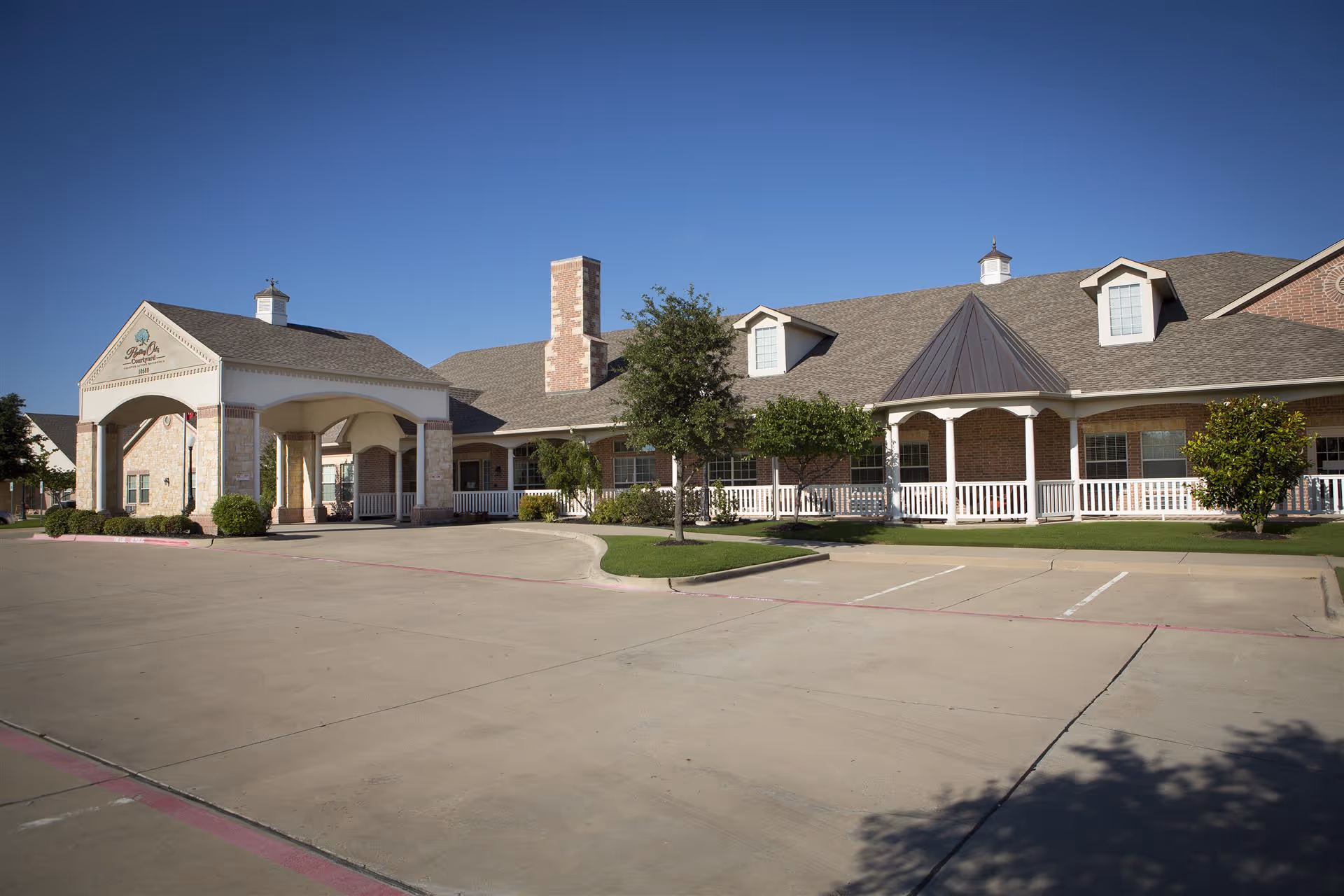 Front exterior of a single-story assisted living building with a covered entrance, white porch railing, landscaped shrubs, and an empty parking lot under a clear blue sky.