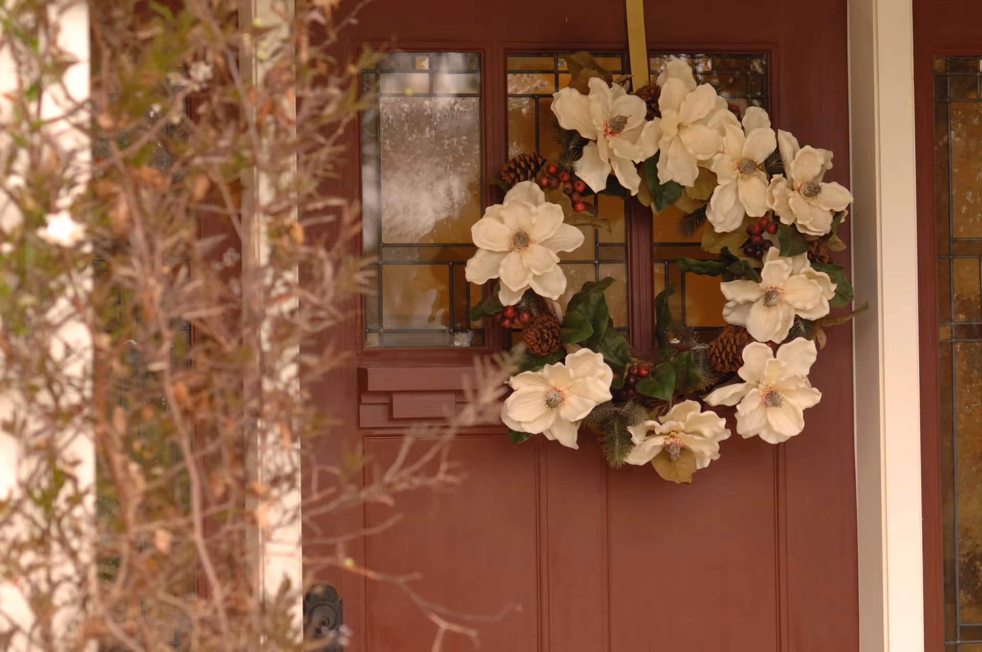 A close-up view of a brown front door decorated with a wreath made of white flowers, pine cones, and green leaves. The door has glass panels with a stained glass design, and some branches with dried leaves are visible in the foreground.