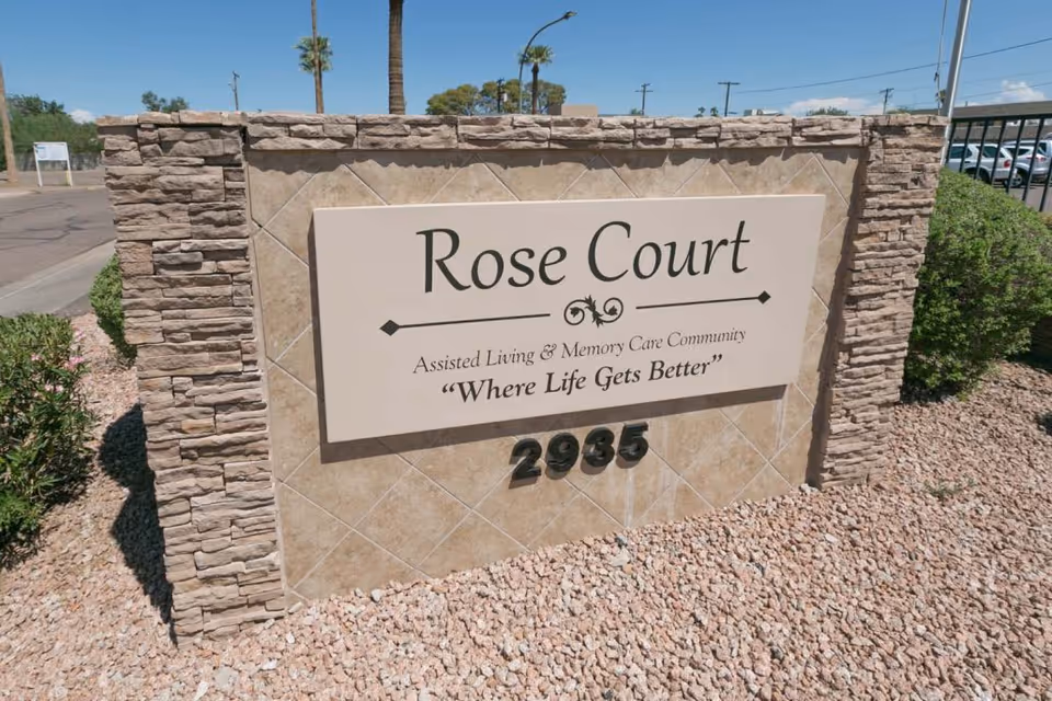 Stone and tile sign for Rose Court Assisted Living & Memory Care Community with the slogan 'Where Life Gets Better' and the address number 2935, surrounded by gravel and some greenery under a clear blue sky.
