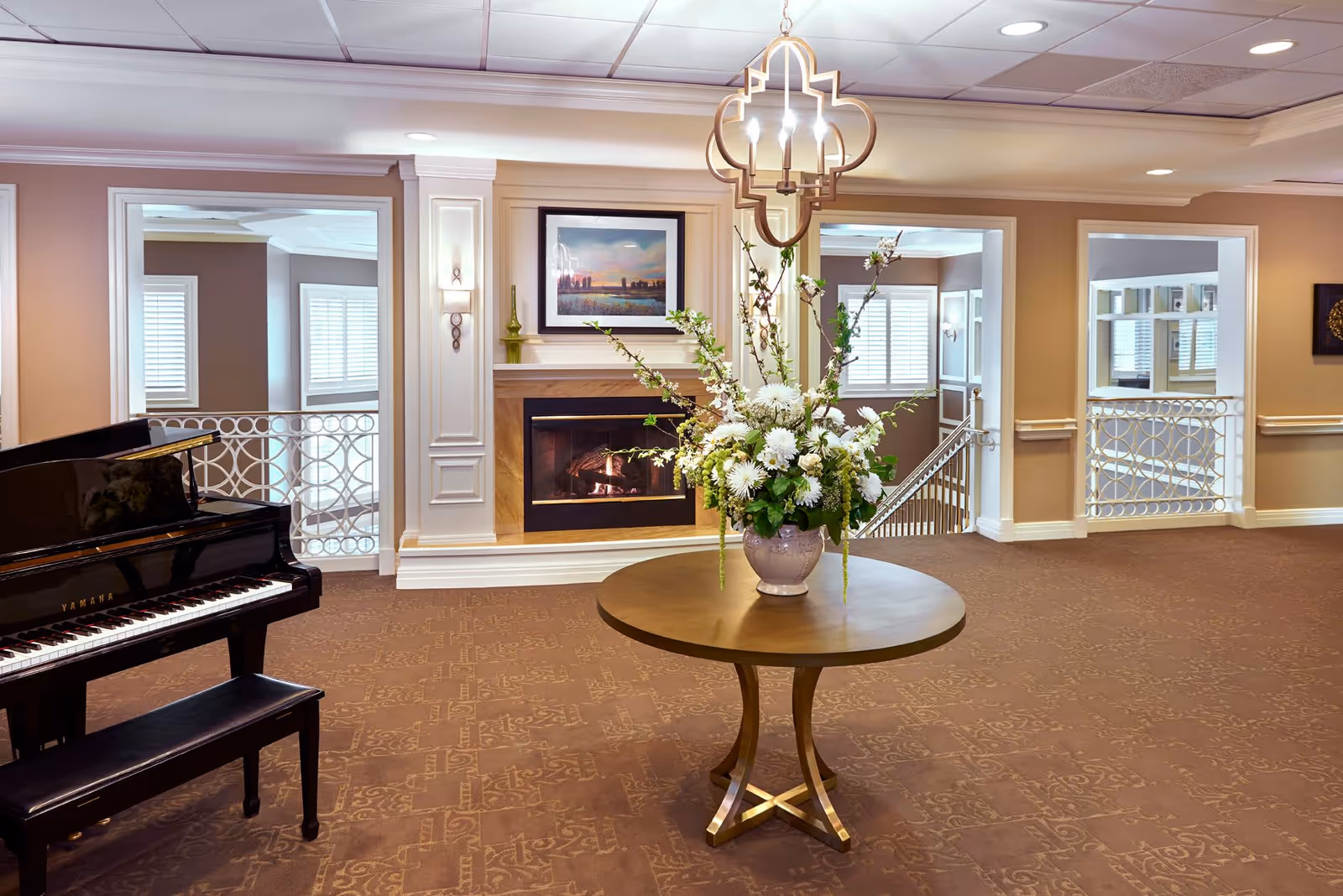 An elegant interior common area featuring a round wooden table with a large floral arrangement in a vase at the center. To the left, there is a black Yamaha grand piano with a matching bench. The background includes a fireplace with a framed picture above it, beige walls with white trim, and two open doorways with decorative railings leading to other parts of the facility. The ceiling has recessed lighting and a decorative hanging light fixture above the table.