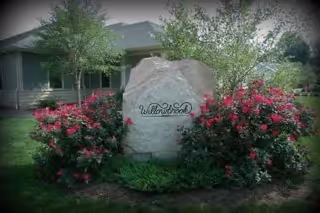 A large landscaping rock engraved with the word "Willowbrook" surrounded by pink flowering shrubs in front of a single-story building.