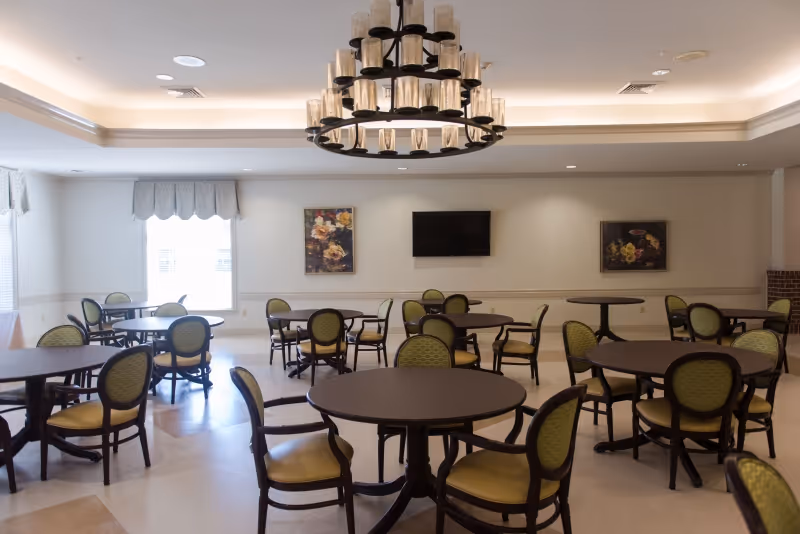 A spacious dining room with multiple round tables and green cushioned chairs arranged neatly. The room features a large chandelier with multiple candle-like lights hanging from the ceiling. There are two framed floral paintings on the wall and a flat-screen TV mounted between them. Large windows with valances allow natural light to brighten the room.