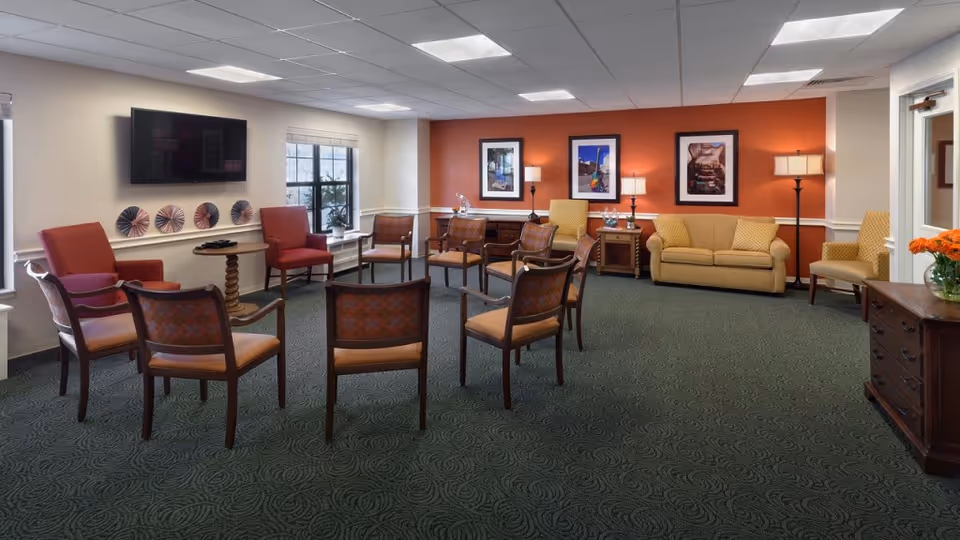 A spacious senior living common area with multiple chairs arranged in a circle on a patterned green carpet. The room has a beige and orange color scheme with framed pictures on the orange accent wall, a beige sofa, side tables with lamps, and a flat-screen TV mounted on the wall. There is a window letting in natural light and a wooden dresser with a vase of orange flowers.