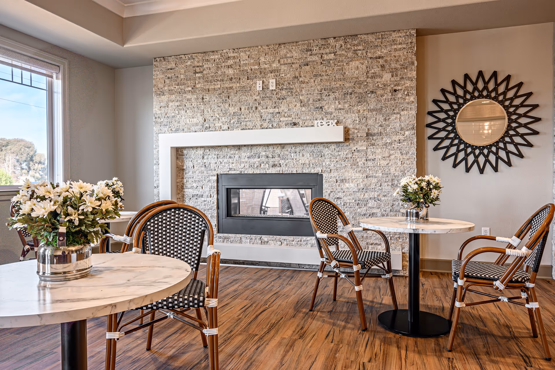 A cozy seating area with two round marble-top tables, each surrounded by two wicker chairs with black and white woven seats and backs. Each table has a silver vase with white flowers. The room features a stone accent wall with a modern rectangular fireplace and a decorative round mirror with a sunburst frame on the adjacent wall. The floor is wood, and there is a large window letting in natural light.