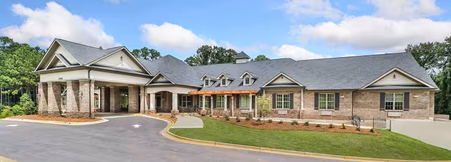 Exterior front view of a single-story senior living facility building with a covered entrance, brick and siding facade, multiple windows, and a paved driveway surrounded by landscaped grass and trees under a partly cloudy sky.