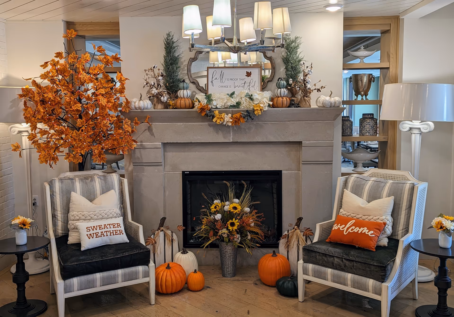 A cozy living room area with a stone fireplace decorated for fall. Two striped armchairs with cushions, one with a 'SWEATER WEATHER' pillow and the other with a 'welcome' pillow, flank the fireplace. The mantel is adorned with small pumpkins, greenery, and a sign that reads 'fall is proof that change is beautiful.' There is a large orange leafy tree on the left, a white floor lamp on the right, and small side tables with flowers beside each chair. Several pumpkins and a floral arrangement are placed in front of the fireplace.