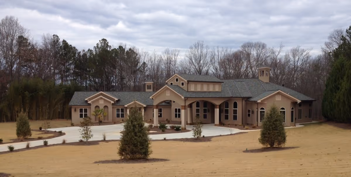 A single-story building with a beige exterior and green roof, surrounded by a lawn with small trees and shrubs. The building has multiple windows and a covered entrance with arches. In the background, there are leafless trees under a cloudy sky.