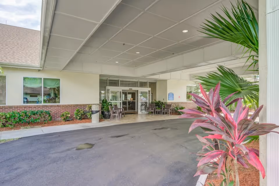 Covered entrance area of Merrill Gardens at Barkley Place with a driveway, potted plants, outdoor chairs, and glass double doors leading inside the building.