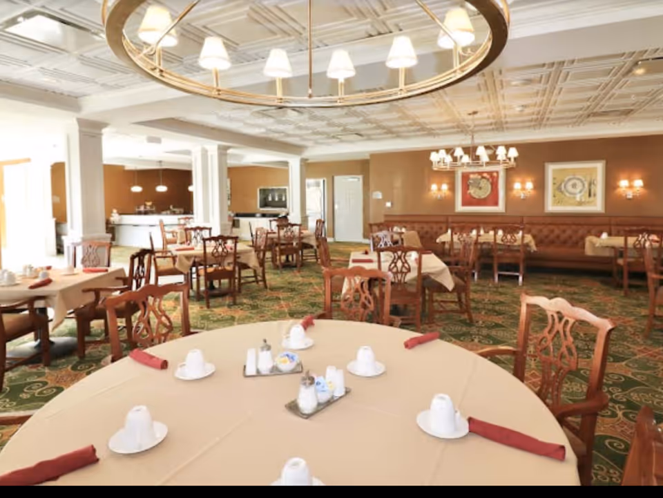 A spacious dining room with round and rectangular tables covered with beige tablecloths, each set with white cups and saucers, red cloth napkins, and condiment trays. The room features wooden chairs with intricate backs, a patterned green carpet, and warm brown walls adorned with framed artwork and wall sconces. A large circular chandelier hangs from the decorative ceiling.