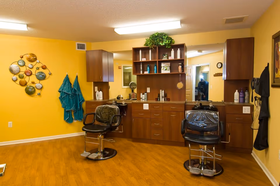 Interior view of a salon area in a senior living facility with two black salon chairs in front of a wooden counter and mirror. The walls are painted yellow, and there are teal towels hanging on the wall along with decorative wall art. Various hair care products are placed on the counter and shelves.
