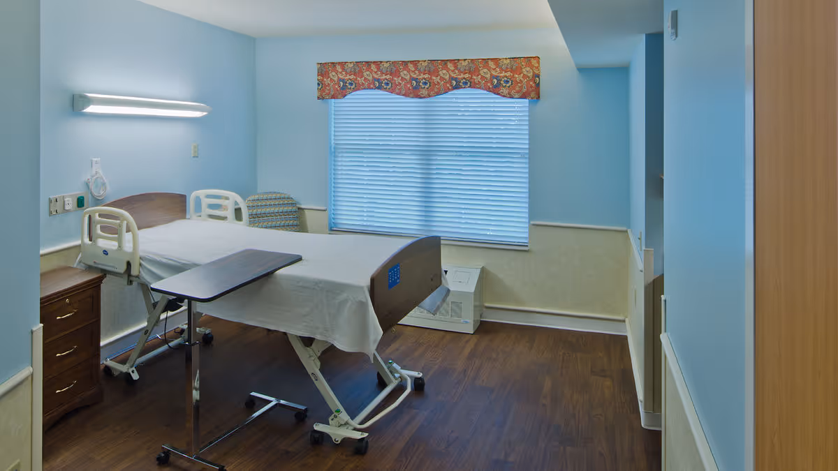 A clean and simple patient room with a hospital bed covered with a white sheet, an adjustable overbed table, a wooden nightstand with drawers, a patterned armchair, and a window with closed blinds and a decorative valance. The walls are painted light blue with a beige wainscoting, and the floor is dark wood.