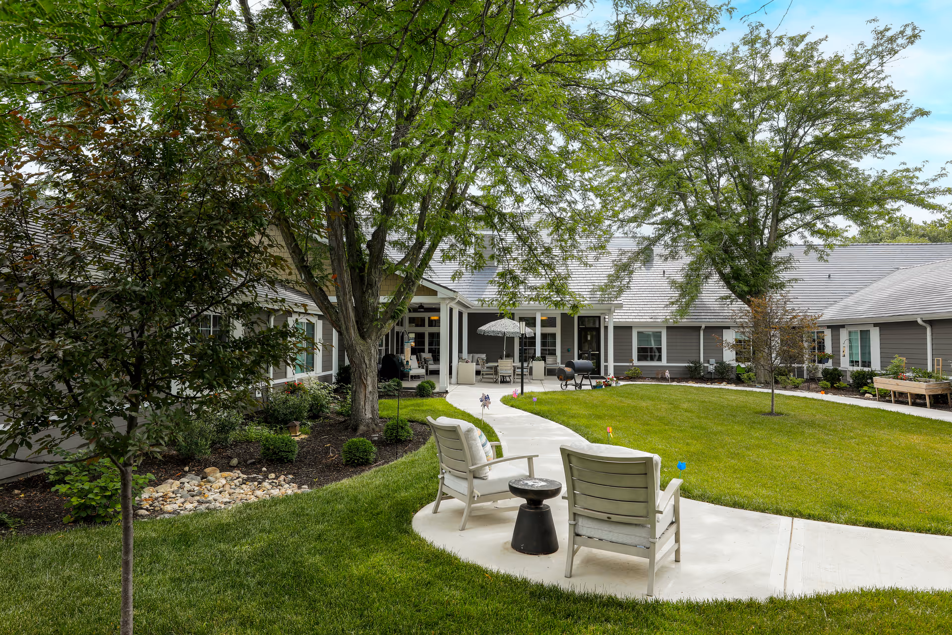 Grassy courtyard at an assisted living facility with two chairs on a curved paved path, trees, and a covered patio area with seating in the background.