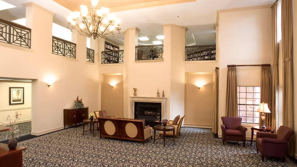 A spacious senior living community lounge with high ceilings, a chandelier, and a fireplace. The room features comfortable seating including a sofa and armchairs arranged around small tables. Large windows with curtains allow natural light to fill the space, and decorative wall sconces provide additional lighting. The carpet has a patterned design, and there is a balcony area overlooking the lounge.