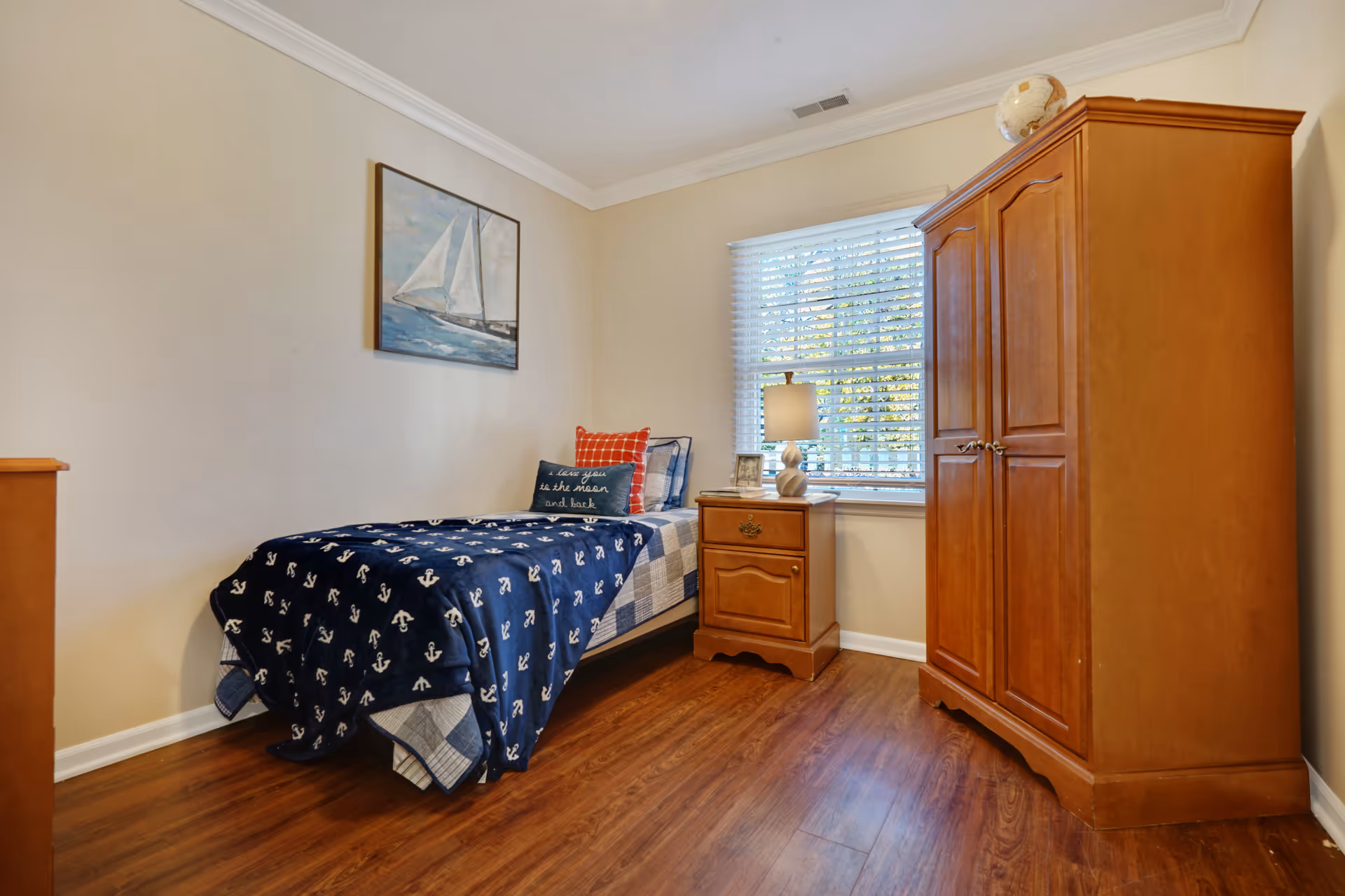 A small bedroom with a single bed covered in a navy blue blanket with white anchors, a red and a navy blue pillow, a wooden nightstand with a lamp and picture frames, a large wooden wardrobe, and a window with white blinds letting in natural light. A painting of a sailboat hangs on the wall above the bed.