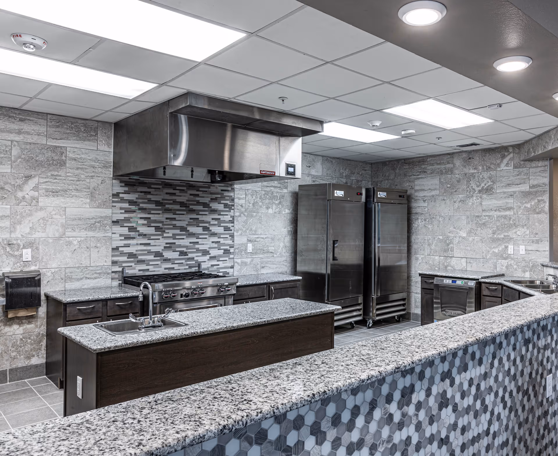 Modern commercial kitchen with stainless steel appliances including two large refrigerators, a stove with a stainless steel hood, granite countertops, a small sink on the island, and a tiled backsplash and walls.