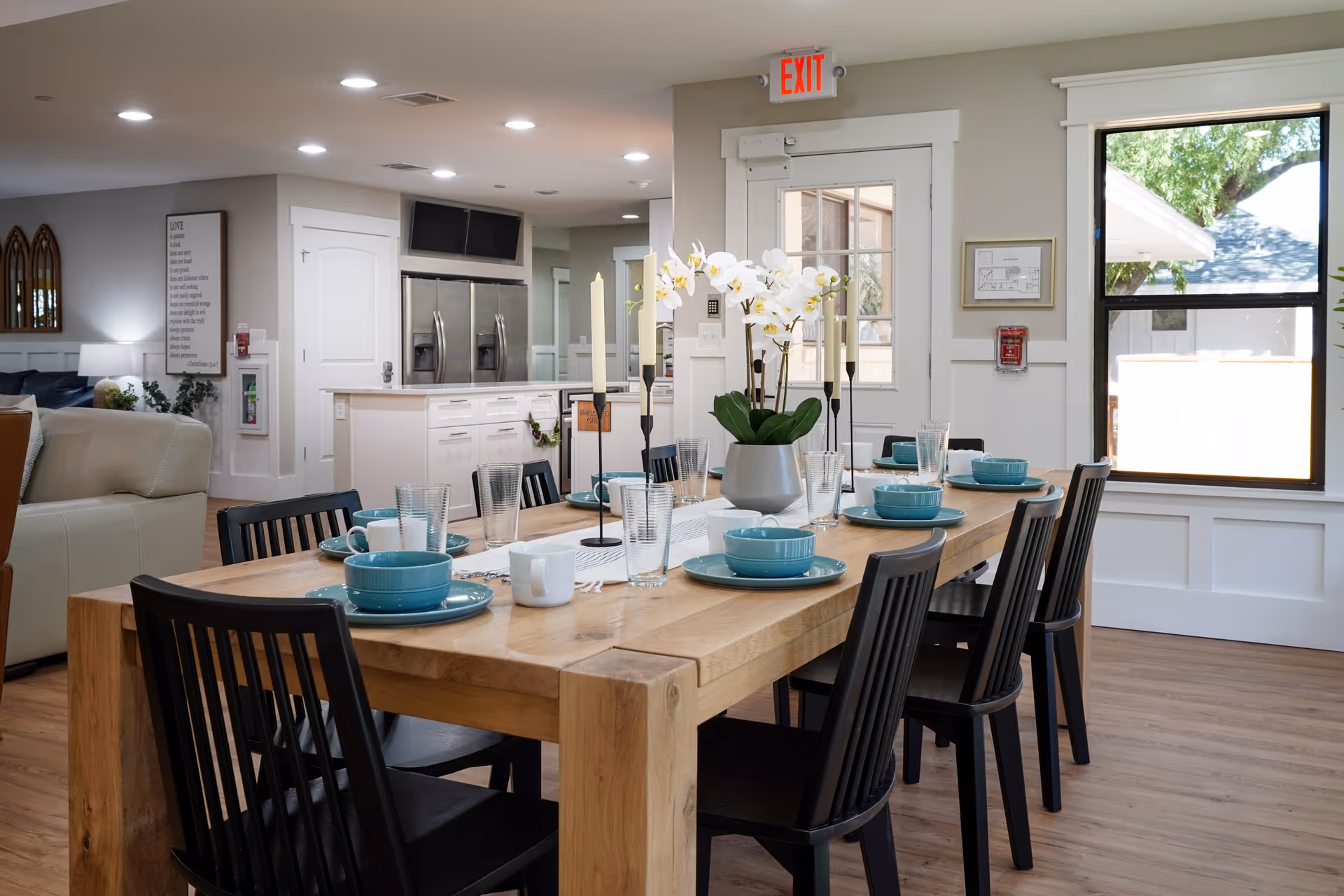 A dining area with a wooden table set for eight people, featuring turquoise bowls, plates, white mugs, and clear glasses. The table is decorated with a white runner, two tall candles, and a vase with white orchids. The room has large windows letting in natural light, a door with an exit sign above it, and a kitchen area with stainless steel appliances in the background.