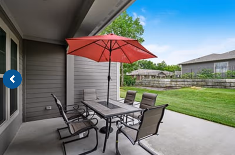 Outdoor patio area with a rectangular table and six chairs arranged around it. A large red umbrella is positioned in the center of the table. The patio is adjacent to a building with gray siding, and a grassy lawn with trees and other buildings is visible in the background under a blue sky.