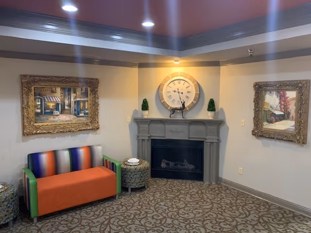 A cozy sitting area with a colorful sofa featuring green armrests and an orange seat cushion, flanked by two patterned ottomans. The walls are decorated with two framed paintings depicting street scenes. A gray fireplace with a large round clock and two small potted plants on the mantel is centered on the back wall. The room has patterned carpet and recessed ceiling lights.