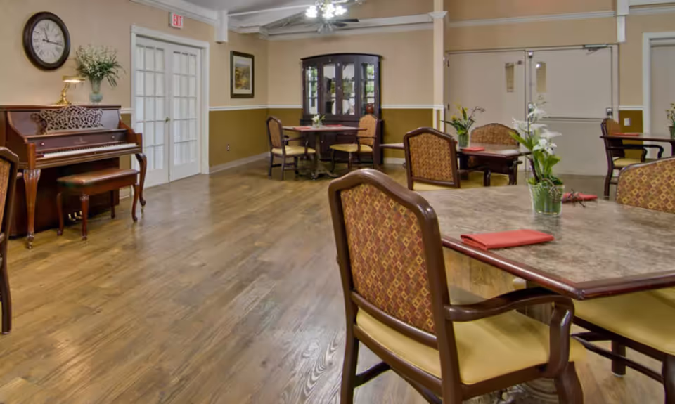 A spacious common area with wooden flooring featuring several tables and chairs arranged for seating. A piano with a bench is placed against one wall, topped with a lamp and a plant. A large clock hangs above the piano. The walls are painted beige with a darker brown lower half, and there are double doors with glass panes and an exit sign above them. The room is decorated with framed artwork and potted plants on the tables.