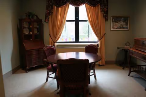 Round wooden dining table with four chairs in a sunlit dining room by a window.